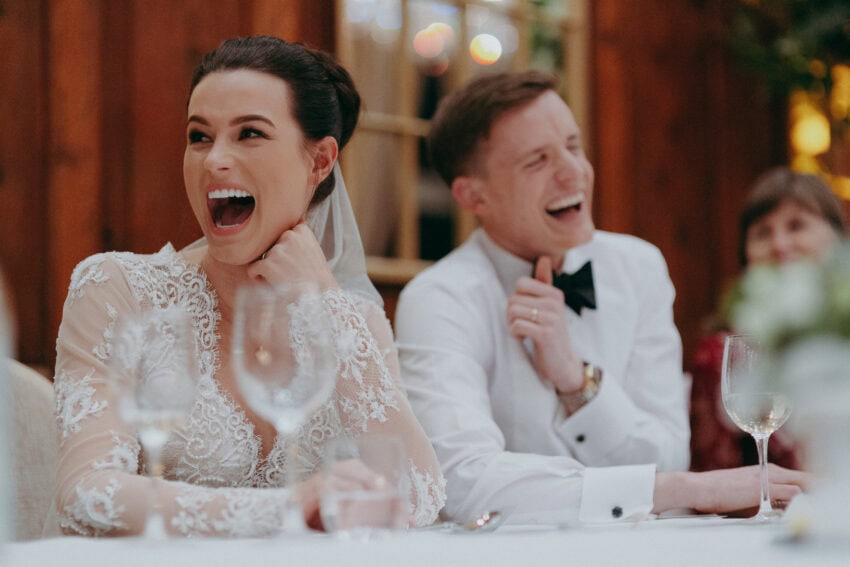 A bride and groom sit at a table, both laughing joyfully. Captured by a wedding photographer, the bride’s lace dress and veil shine softly beside the groom in his white shirt and bow tie, with warm lighting in the blurred background.