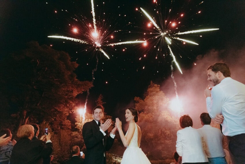 A bride and groom stand outdoors at night, smiling and clapping as bright fireworks light up the sky. Guests and a wedding photographer capture the magical moment, with trees visible in the background.