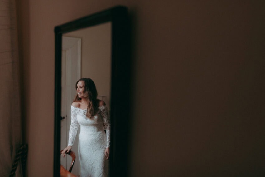 A woman in a white off-the-shoulder lace wedding dress is reflected in a mirror, standing beside a chair and smiling softly in a softly lit room, captured beautifully by a Creative Wedding Photographer.