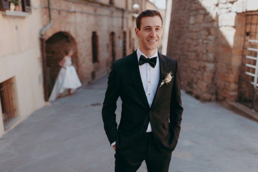A smiling man in a black tuxedo stands outdoors on a stone street, hands in pockets, with a boutonnière on his lapel. Captured by a wedding photographer, a woman in a white dress walks away in the softly blurred background.