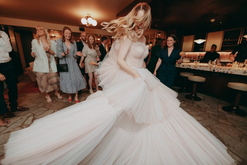 A bride in a voluminous white gown dances energetically at a wedding reception, as a Wedding Photographer captures the joyful moment amid smiling guests in semi-formal attire inside a warmly lit venue.