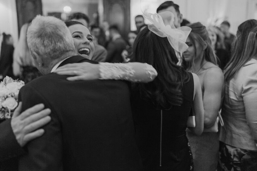A bride laughs joyfully as she hugs a man in a suit at a crowded, elegant indoor event. Other guests, dressed formally, mingle in the background. Captured by a wedding photographer, the image is in black and white.