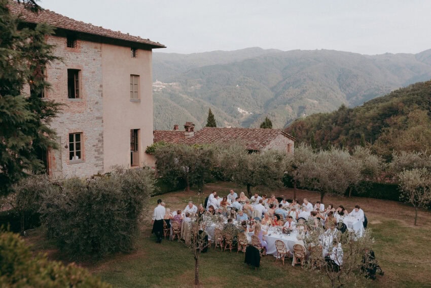 A large group of people dine outdoors at round tables in a garden beside a stone building, surrounded by trees and mountains—a perfect scene for a Creative Wedding Photographer to capture unforgettable moments.