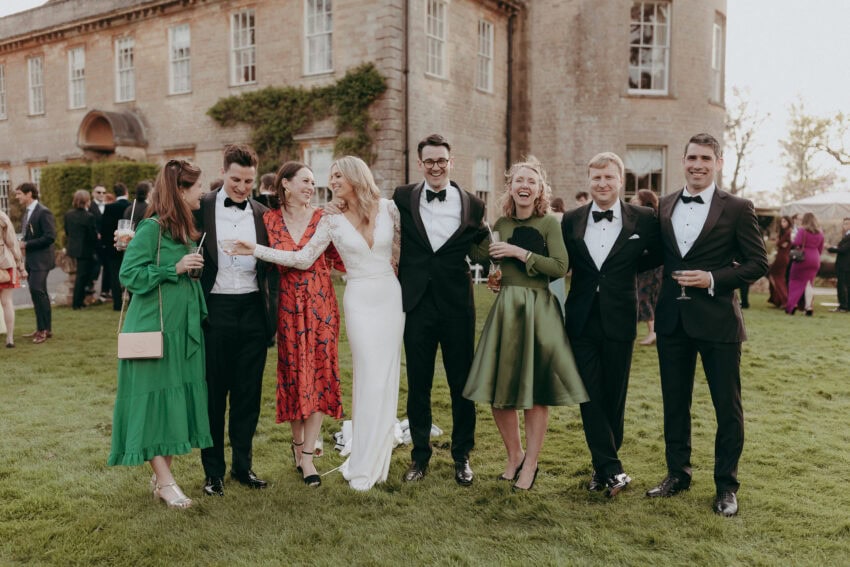 A group of seven people dressed in formal attire stand on grass, smiling and laughing together in front of a large, elegant stone building at an outdoor event, expertly captured by a wedding photographer.