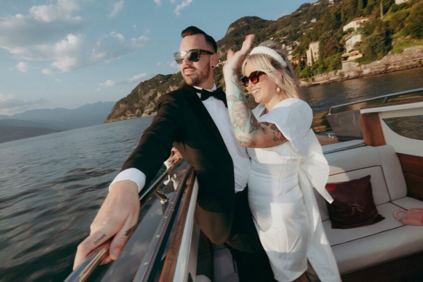 A couple dressed in formal attire, the man in a tuxedo and the woman in a white dress with sunglasses, smile and pose on a boat against a scenic lakeside and mountain backdrop under a partly cloudy sky, captured by a creative wedding photographer.
