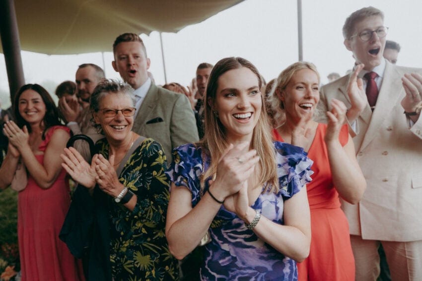 A group of well-dressed people stand closely together outdoors, smiling and clapping enthusiastically. Captured by a creative wedding photographer, the joyful, festive scene shows men and women celebrating together under a canopy.