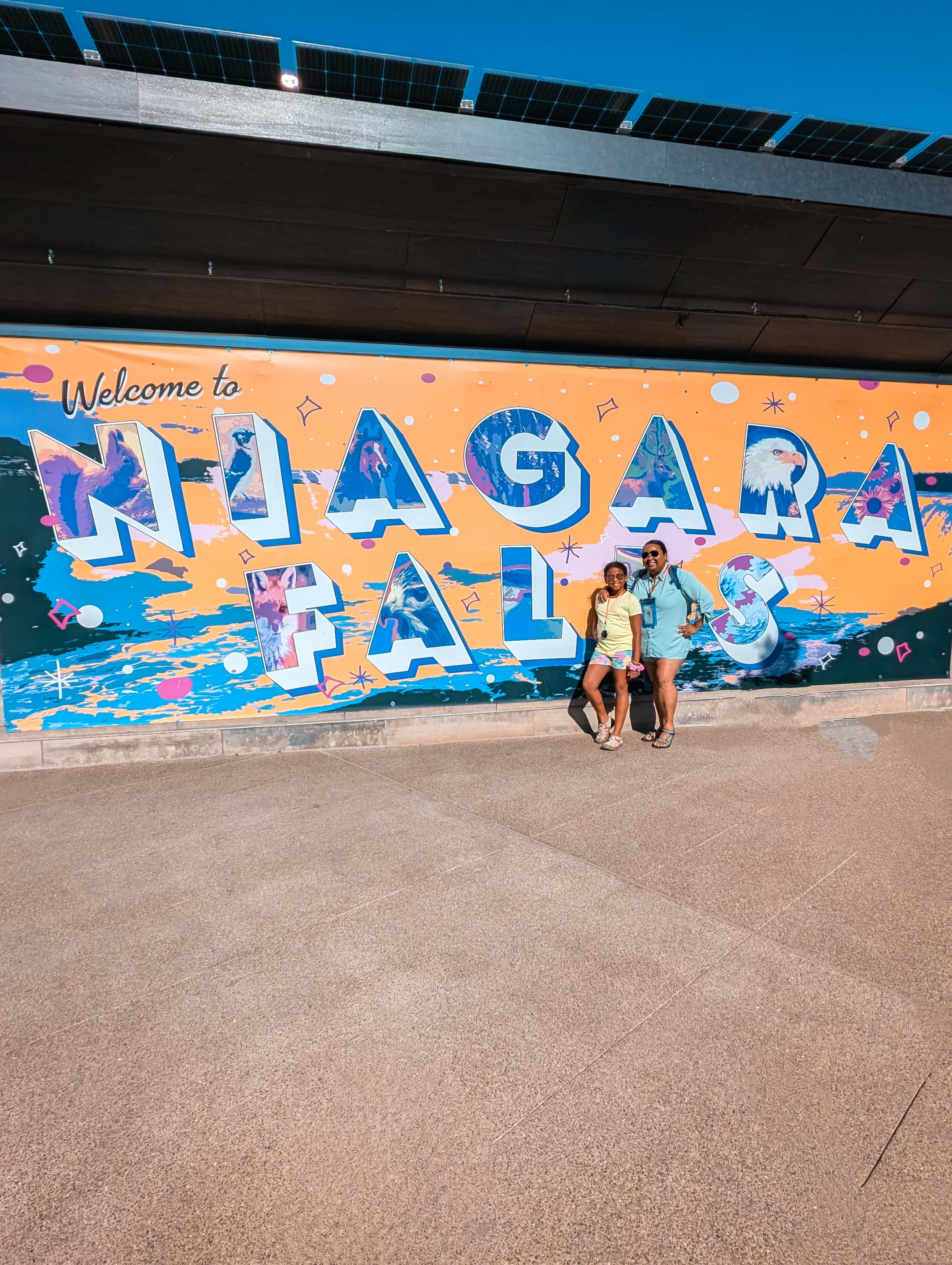 Mom and daughter posing in front of a mural that says “Niagara Falls” 