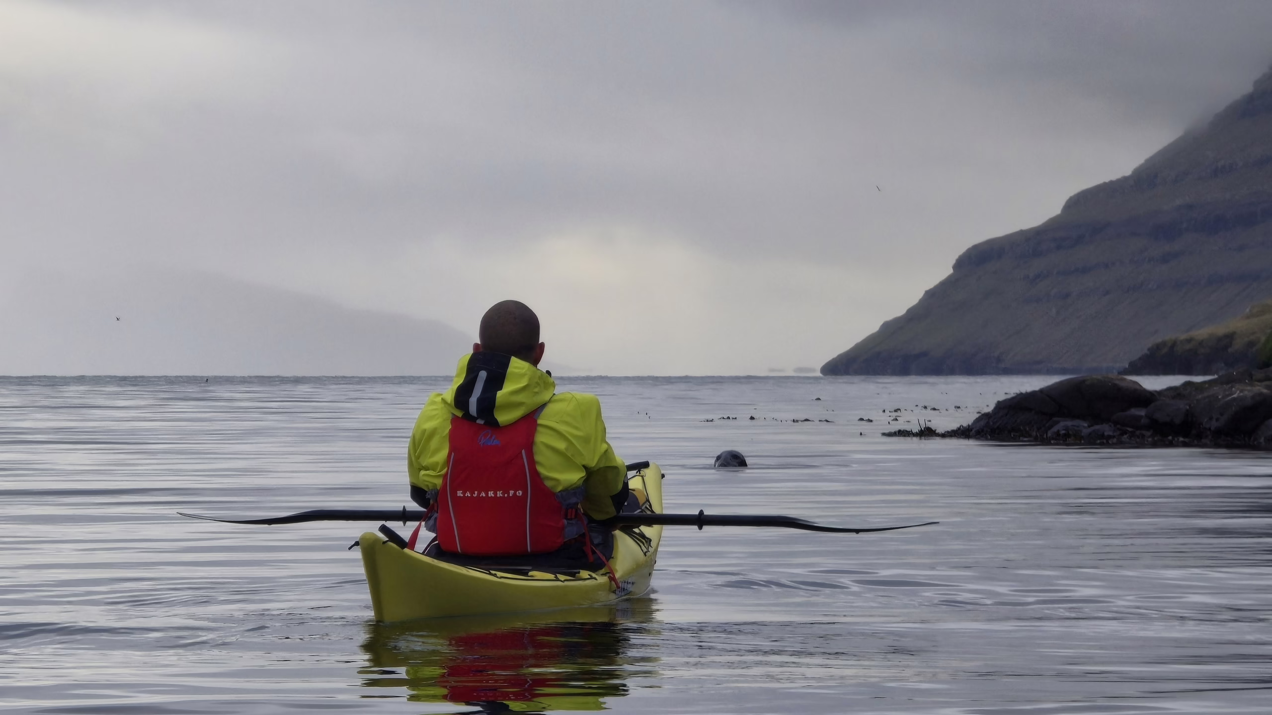 paddling a bright yellow kayak on calm water, facing a rocky coastline of the Faroe Islands under cloudy skies