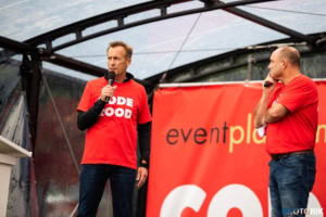 Broadcaster speaking at an outdoor event, with a "rode goed" T-shirt, holding a microphone, addressing an audience at a public gathering.