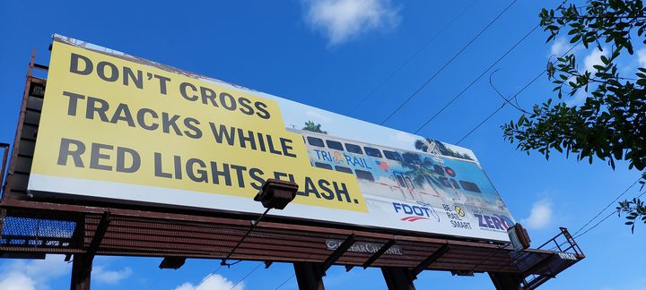 Roadside billboard reading Don’t cross tracks while red lights flash, shown against a bright blue sky.