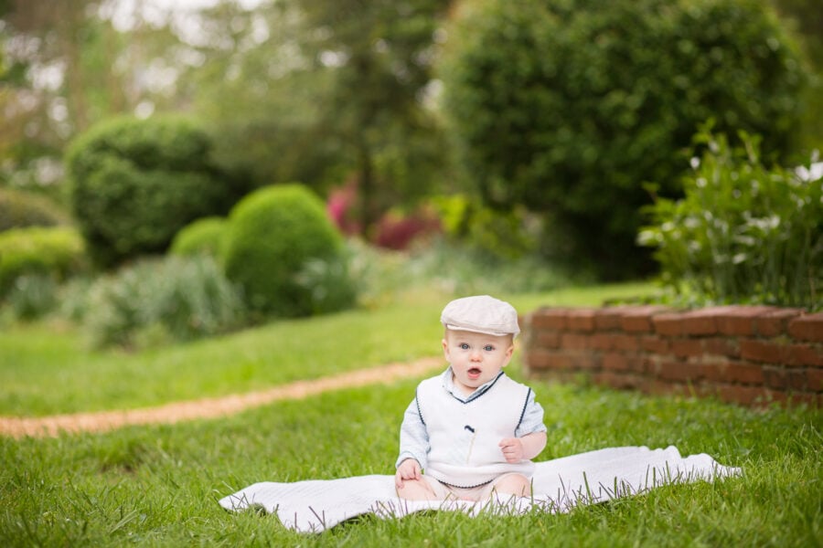 Six-month-old boy wearing a cap sits on a blanket in the grass.