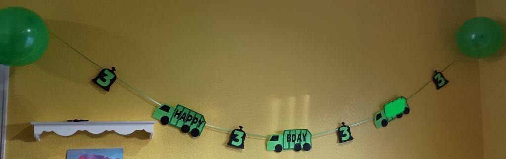 DIY garbage truck banner. There are trash trucks made of paper alternating with garbage bags with 3s on them. On both ends there is a green balloon