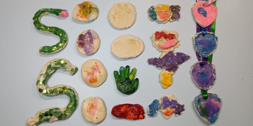 Picture of several things made by a toddler and a preschooler out of a salt dough recipe. There are S shaped snakes covered in sequins, painted disks, baby feet and hand prints, baby feet and hand crayons, pups, a mermaid, and pup badges.