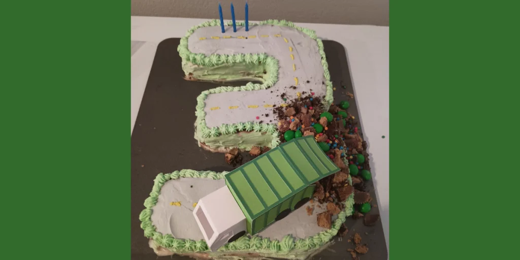 Picture of a three shaped garbage truck cake. The top looks like a road with a paper trash truck on top with crunched up candies coming out the back of it. There are three blue candles in the top