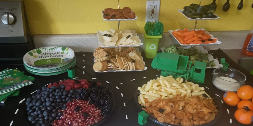 Picture of kid's party food set out on a black tablecloth painted to look like a road. A trash truck toy is on the road and several fake flies are on the food