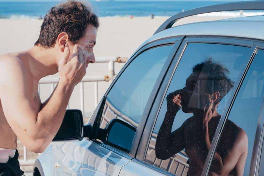 A man putting on sunscreen in his car window reflexion before hitting the waves. The best sunscreen for surfers is reef safe.