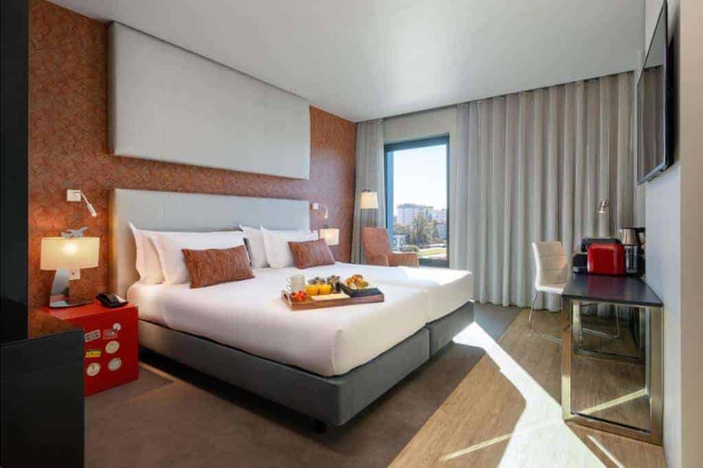 A stylish hotel room featuring two twin beds with crisp white linens, decorative pillows, and a contemporary orange accent wall with artistic portraits. A desk, chair, and coffee station are situated by the window, offering a cozy yet modern atmosphere. This Lisbon airport hotel with free shuttle offers a comfortable stay for travelers seeking both style and convenience.