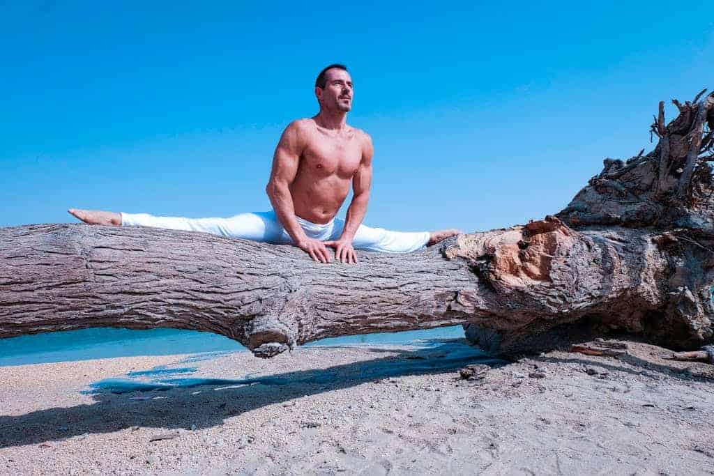 A fit man performs yoga splits on a beach log under a clear blue sky, showcasing flexibility and balance.