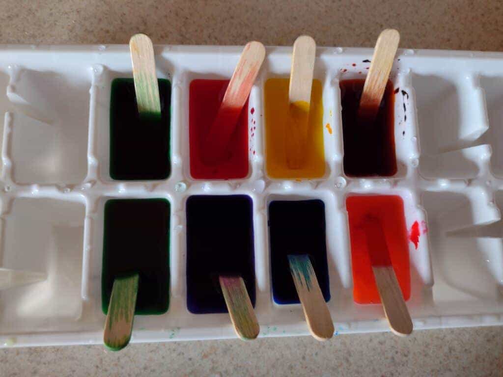 Picture of food coloring mixed with water in an ice cube tray. Short popsicle sticks are sticking out of each one
