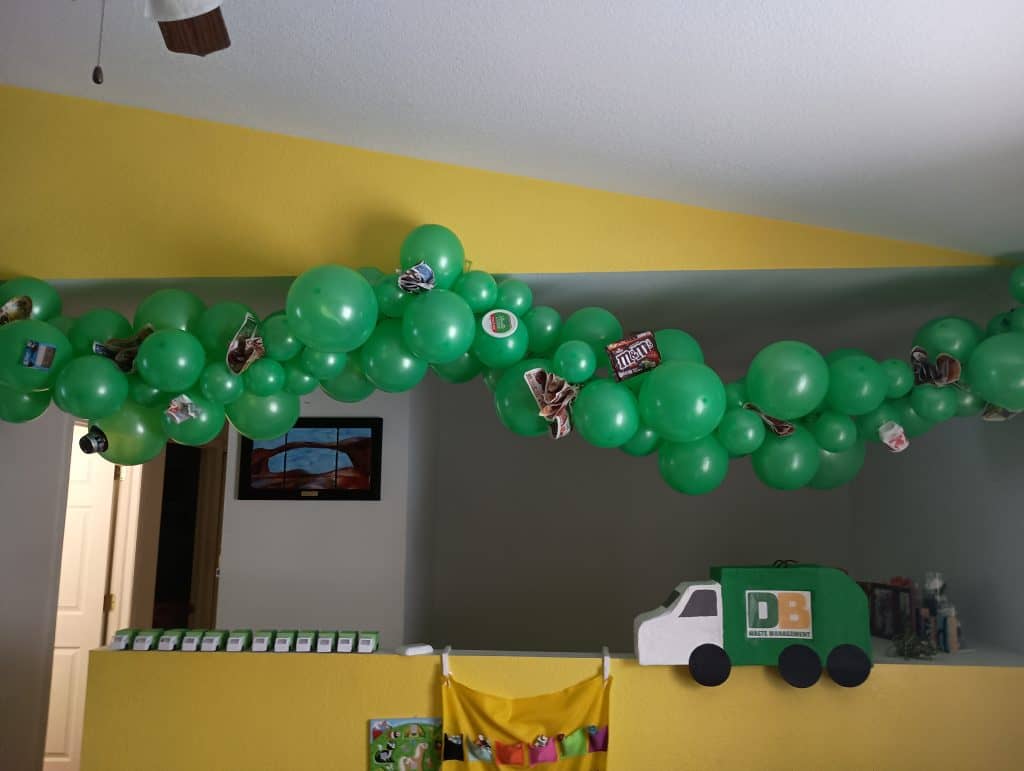 Picture of a green balloon garland hanging from the ceiling. There is trash on the garland for a garbage truck birthday party