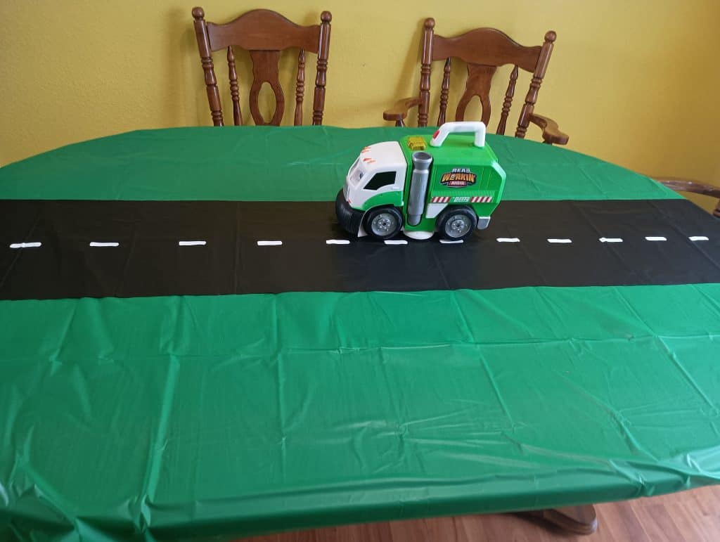 Picture of a dining table covered in a green tablecloth with a black road down the middle and a trash truck toy on the road