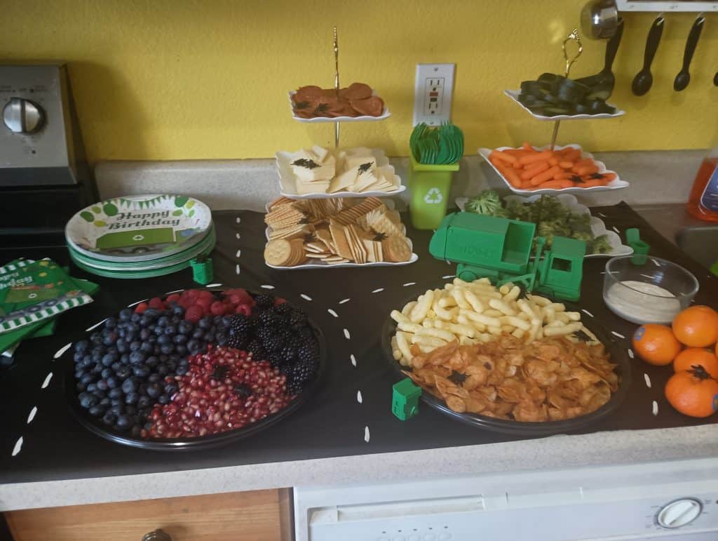 Picture of fruit, veggies, chips, and cheese and crackers on a tablecloth painted with a road. A 3d printed garbage truck is on the road along with a few mini plastic trash cans
