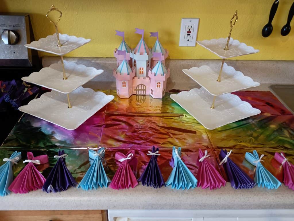 Picture of the food area for a princess party. There is a paper castle in the middle with tired trays on both sides of it. At the front is a line of blue, pink, and purple napkins folded to look like dresses