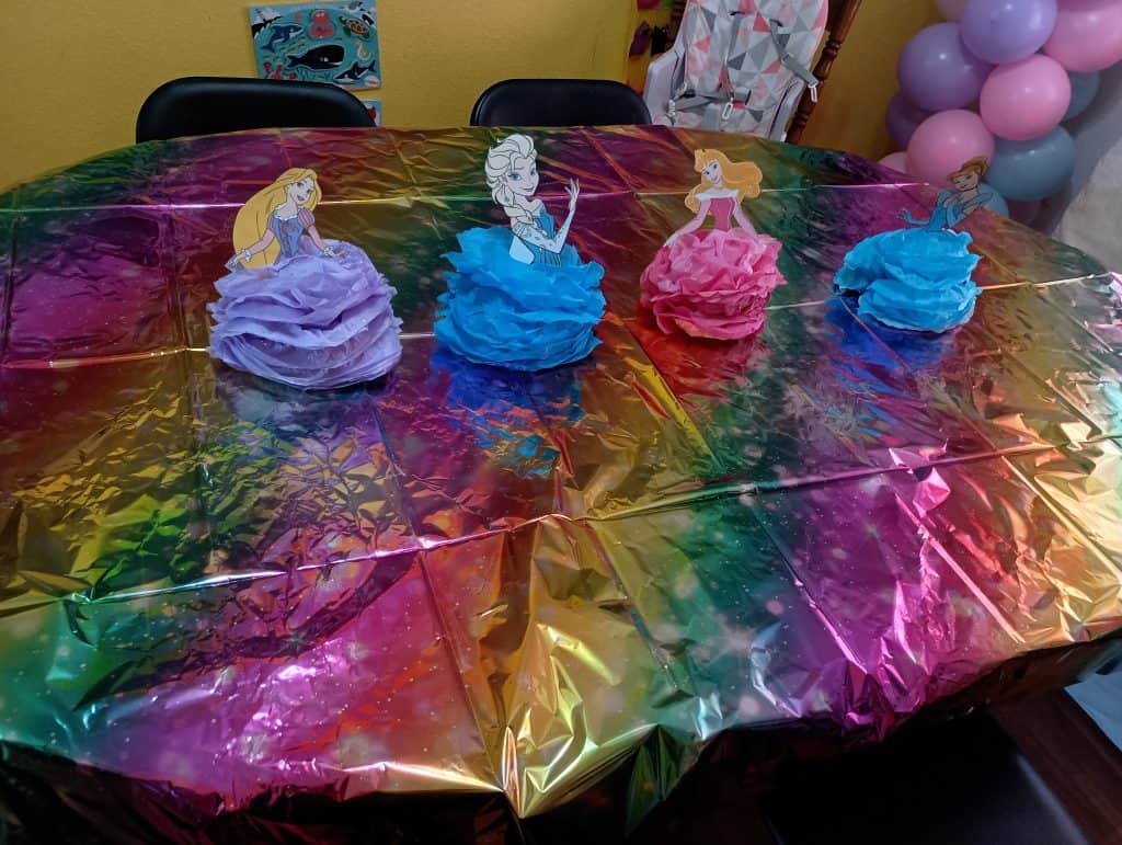 Picture of a table covered in a shiny rainbow tablecloth and four princess decorations. Each decoration is made of a tissue paper pom pom as the princess' dress and a laminated picture of their torso and head