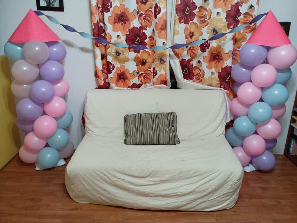 Picture of two balloon pillars on either side of a couch. The balloons are pink, purple, and blue. There is a pink cone on the top of each balloon pillar and streamers go between the tops of them