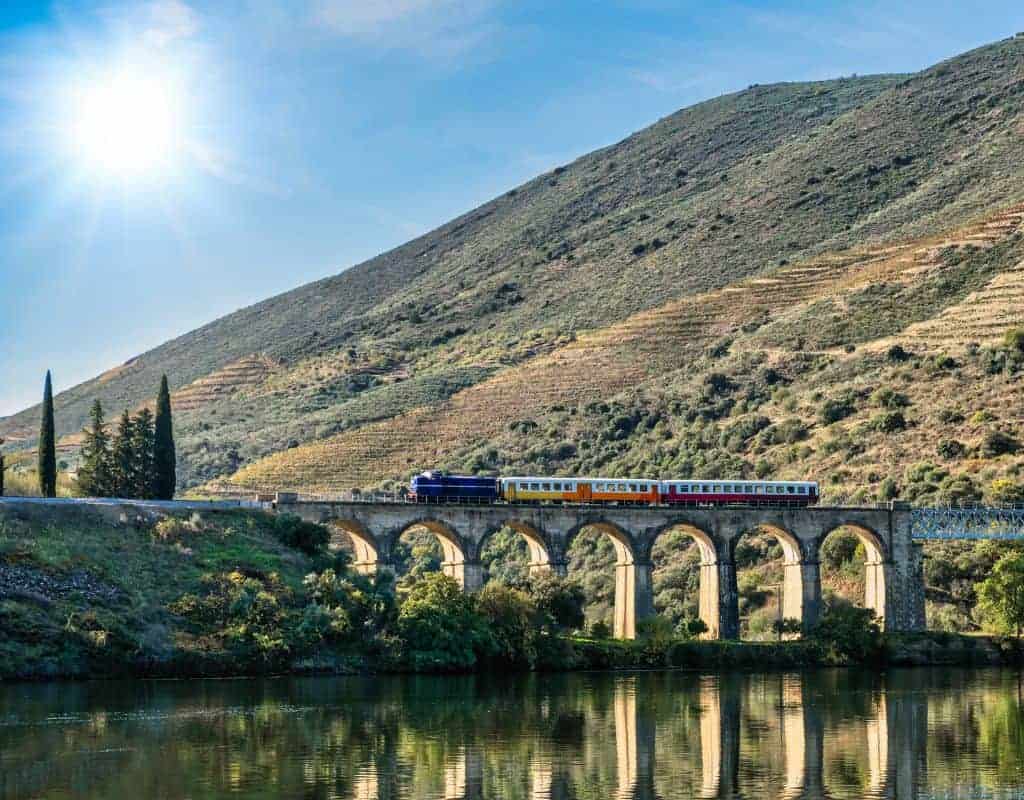 A train travels over an arched bridge against the backdrop of the Douro Valley's terraced vineyards and blue sky, symbolizing a scenic journey through one of Portugal's most beautiful wine regions.