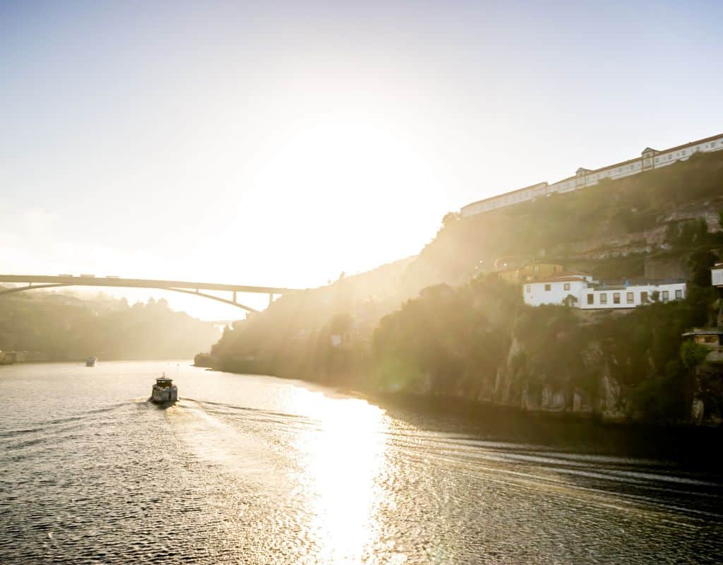 The sun sets over the Douro River, casting a warm glow over the landscape with a boat in the foreground and a distant bridge, illustrating a tranquil moment in the valley. This is the scene you get to see on your return from your trip from Porto to Douro Valley by train.