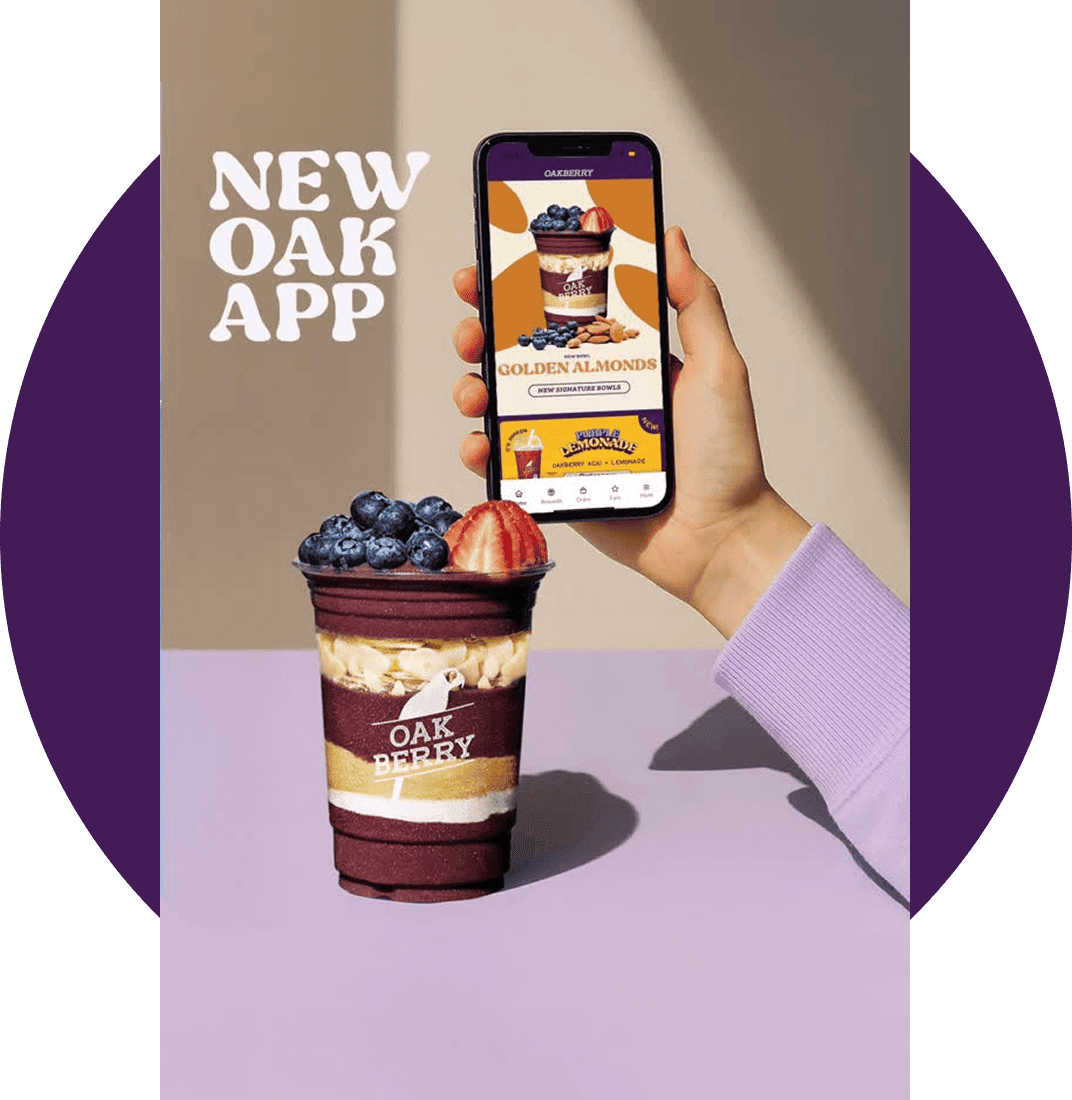 Promotional image for the new Oakberry app, featuring a hand holding a phone displaying the app interface and a branded açaí bowl topped with blueberries and strawberries.