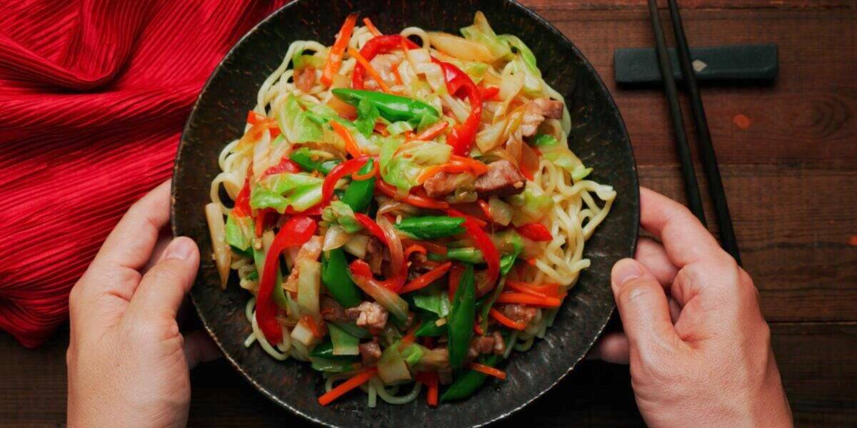 Chop Suey in a dark plate being placed on a dark wood table with a red napkin and chopsticks.