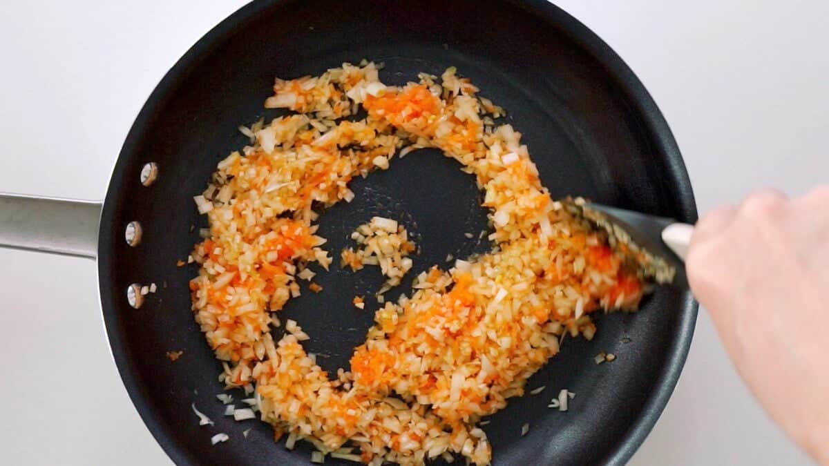 Stirring onions, garlic and grated carrots together in a frying pan.