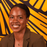 A woman with dark hair pulled back wearing a pendent necklace and brown blazer.