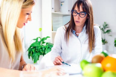 A Certified Nutrition Specialist with a female patient in her office with fruits and explain how she became a certified nutrition specialist