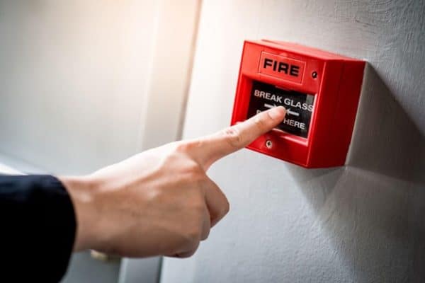 A man pointing at a red fire alarm switch on a concrete wall in an office building.