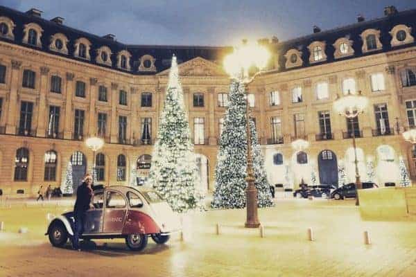 A man in a santa hat poses with his classes citroen in front of an elegant paris facade and its lit up christmas trees.