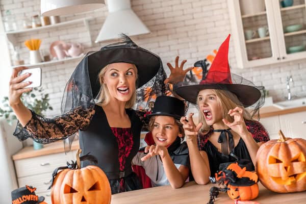 Three people in witch costumes take a selfie at a Halloween-themed kitchen table with carved pumpkins nearby.