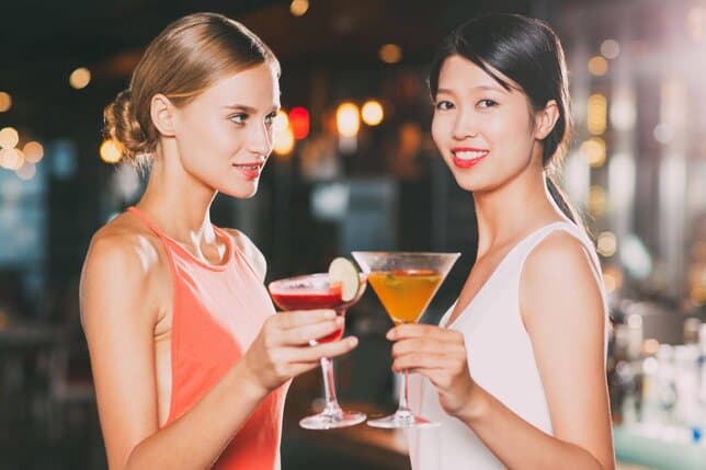 Two women in a bar toasting with colorful cocktails, smiling at the camera.