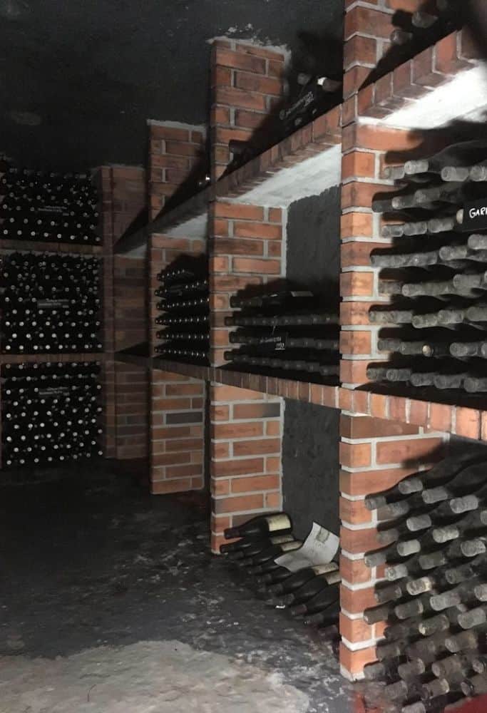 An underground wine cellar with rustic brick shelving filled with numerous bottles of wine. The darkness and shadows emphasize the cool, controlled environment for wine preservation.
