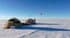 Polar camping scene: two tents on snow, gear scattered, a person in a red jacket near equipment, with a red paraglider overhead in blue sky.