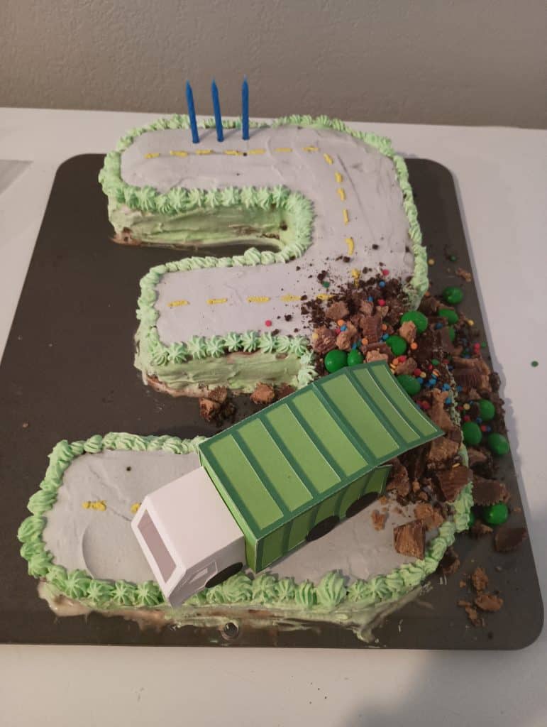 Three shaped birthday cake. There is a frosting road on top with a paper trash truck and crunched up candies pouring out
