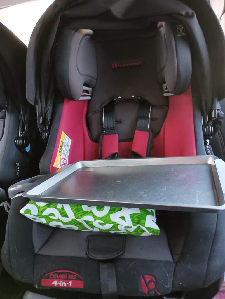 Picture of a DIY car seat tray made of a cookie sheet and a green number pillow attached to a car seat
