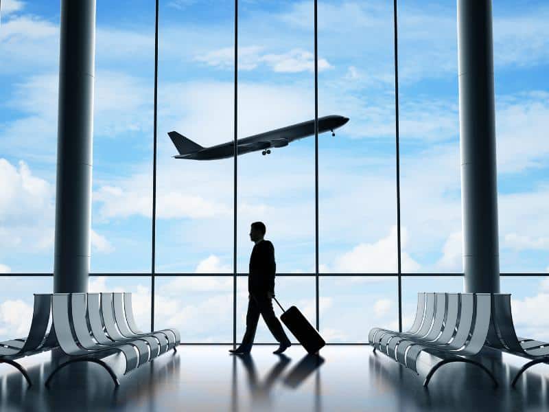 Shadow of a man walking with his suitcase in an airport lounge with a floor to ceiling window in the background and a plane taking off in the sky on a sunny day.
