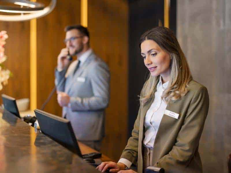 A woman and a man assisting costumers at reception in a hotel. Both are wearing pant suits and are happy to serve customers who might be arriving from the airport on a shuttle.