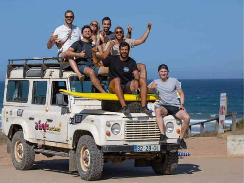 A group of surfers sitting on the top and hood of a white land rover with a surf board and the sea in the background.