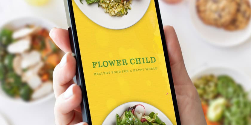 Mockup of hand holding a mobile phone showing the Flower Child mobile app above a blurred background of Flower Child dishes