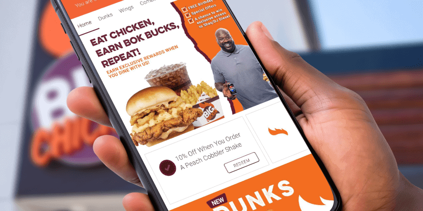 Close-up of a hand holding a smartphone displaying the Big Chicken restaurant app. The screen shows a promotional offer featuring Shaquille O’Neal, with the headline ‘Eat Chicken, Earn Bok Bucks, Repeat!’ and a 10% discount on a Peach Cobbler Shake. The background includes the blurred Big Chicken restaurant sign.