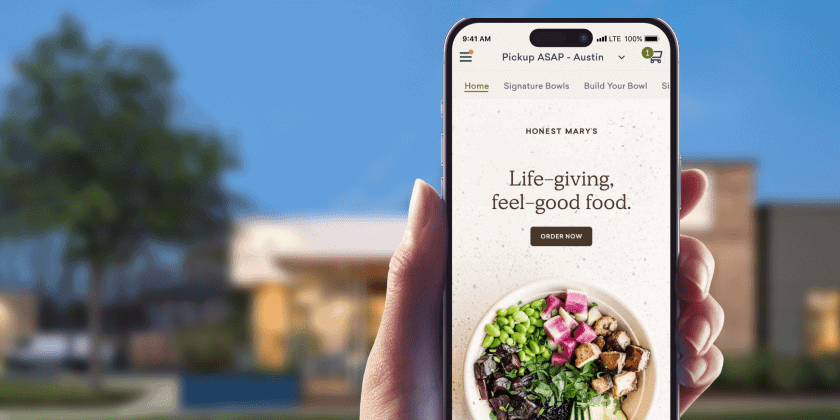 A hand holding a smartphone displaying Honest Mary’s mobile website, showing a bowl of colorful fresh ingredients and the text ‘Life-giving, feel-good food’ with an order button, against a blurred outdoor background.